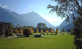 A wide green meadow surrounded by mountains and trees. In the background, autumn-colored trees and a clear sky can be seen.