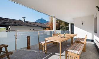 A modern balcony with a wooden table and benches. In the background, mountains and a clear blue sky can be seen.