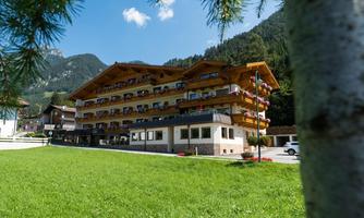 A charming hotel building with wooden cladding, surrounded by a green meadow and mountains in the background. The sky is blue with a few clouds.