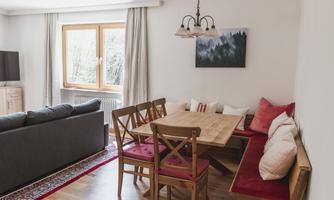 A cozy living space with a sofa and a dining table. Cushions hang on the wall and there is a large window with a view outside.