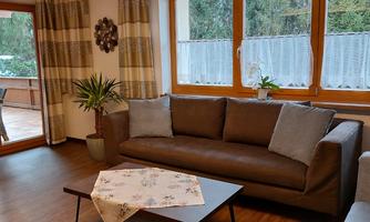 A cozy living room with a large sofa and a table.  
Lots of daylight streams through the windows and there are plants in the room.