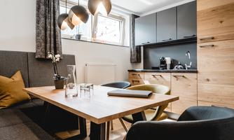 A modern living and dining room with a wooden table and comfortable chairs. The kitchen in the background is well-equipped and stylishly furnished.