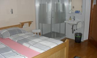 A small room with a double bed and a shower.  
There is a sink and a trash can in the corner.