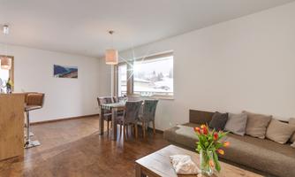 A modern living room with a cozy sofa and a wooden table. There is a dining area with chairs and large windows that provide plenty of light.