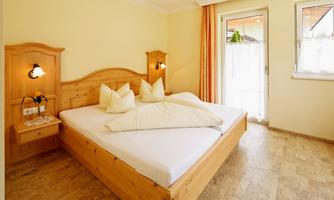 A cozy bedroom with a large double bed and bright wooden furniture. Large windows provide plenty of daylight and a view outside.