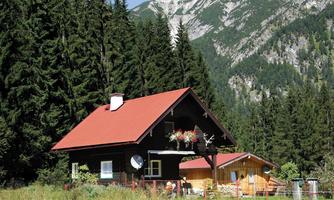 A cozy wooden house with a red roof, surrounded by tall fir trees. In the background, majestic mountains rise under a clear sky.