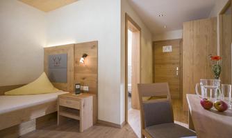 A bright room with wood decor that includes a single bed, a table, and a chair. On the table are apples and a water carafe.