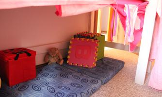 A cozy play area under a bed with a blue mat, a plush teddy bear, and colorful building blocks. The environment is child-friendly and inviting.