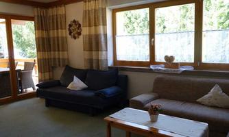 A cozy living room with a sofa and an armchair. Large windows let in plenty of daylight and offer a view outside.