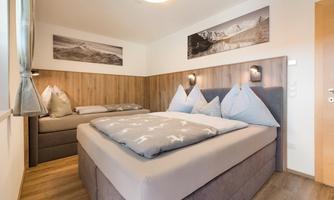 A modern bedroom with a double bed and two pillows. Pictures of mountains hang on the walls, and the room has a warm, inviting wooden look.