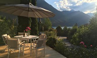 An inviting outdoor area with a table and chairs under a sunshade. In the background, mountains and a calm lake can be seen.