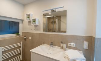 A modern bathroom with a sink, a mirror, and decorative shelves. The walls are in neutral colors, and there is a window for natural light.