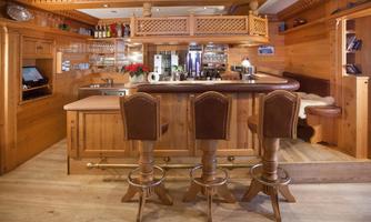 A cozy wooden bar with three bar stools and an inviting atmosphere. Shelves with drinks and a comfortable seating area decorate the space.