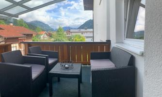 A cozy terrace with rattan furniture and a table. In the background, mountains and a clear sky can be seen.
