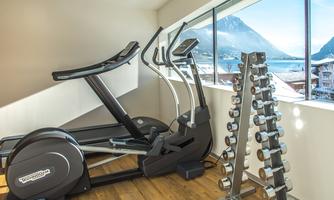 A modern gym with a cross trainer and a barbell bench.  
Large windows offer a view of the mountains and the lake.