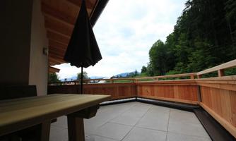 A cozy terrace with wooden planks and a sun umbrella. In the background, there are green trees and a cloudy sky.