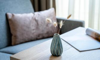 A stylish vase with dried flowers stands on a wooden table. In the background, there is a gray sofa and a window with bright light.