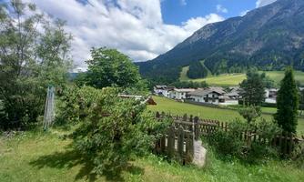 A picturesque landscape with green meadows and a view of mountains. In the background, small houses and trees can be seen under a blue sky with clouds.