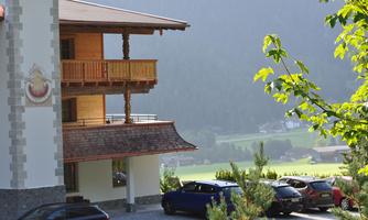 An elegant building with a wooden balcony and a beautiful view of the surrounding mountains. Several cars are parked in front of the building on a paved parking lot.
