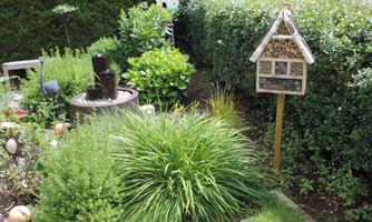 A well-maintained garden with green plants and a decorative insect hotel. In the background, a small fountain and a birdhouse can be seen.