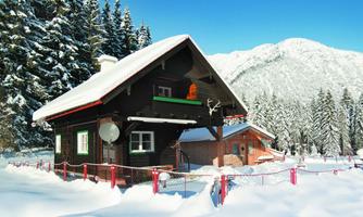 A cozy holiday cabin in the snow, surrounded by fir trees and mountains. The clear blue sky creates a wintry atmosphere.