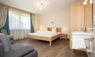 A modern bedroom with a large bed and a cozy seating area. The walls are bright, and the windows are flooded with light.