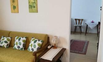 A cozy living room with a green sofa and colorful cushions. In the background, a dining table is visible.