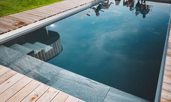 A modern swimming pool with clear water and a wooden deck. The surroundings are green and inviting.
