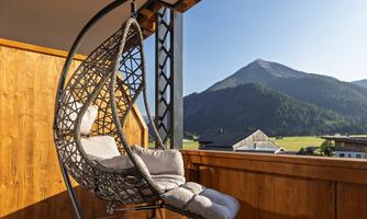 A cozy hanging chair on a terrace with a view of the mountains. The sky is clear and the surroundings are green and natural.