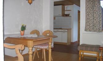 A simple, cozy dining room with a wooden chair and table. In the background, a small kitchen with white cabinets and tiles can be seen.