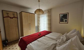 A cozy bedroom with a large double bed and a red bedspread. There is a wardrobe and a window that lets in plenty of daylight.