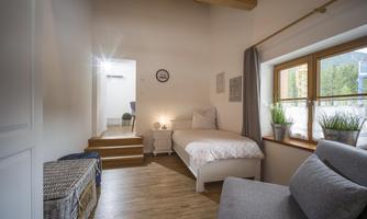 A cozy room with a single bed and a gray armchair.  
Large windows let in plenty of light and plants create a fresh atmosphere.