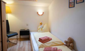 A cozy room with a single bed, a chair, and a lamp. Colorful towels and a plush bear create an inviting atmosphere.