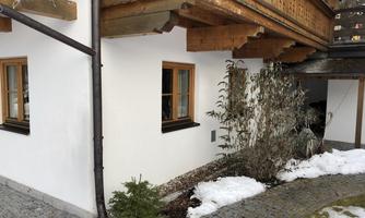 A modern house with wooden decorations and large windows. Snow is lying in front of the house and there is some greenery in the foreground.