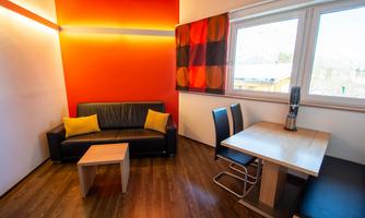A modern living room with a black sofa and colorful cushions. A wooden table and chairs complement the space, which is characterized by a warm wall color.