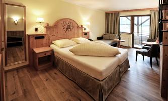 A cozy hotel room with a double bed and wooden decor. A seating area and large windows provide plenty of daylight.