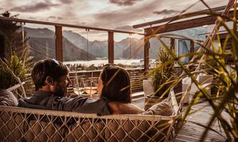 A couple sits comfortably in a hammock overlooking a lake and the mountains. The evening mood creates a romantic atmosphere.