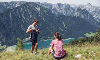 A couple is sitting on a mountain overlooking a clear lake and the surrounding mountains. The atmosphere is relaxed and sporty.