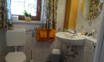 A bright bathroom with a toilet, a sink, and a shower. The windows are adorned with floral curtains, and there are orange towels.