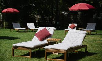 Two loungers on a green meadow with red sun umbrellas in the background. The loungers are equipped with white cushions and red pillows.