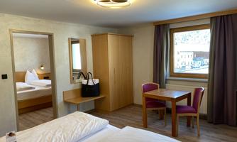 A modern hotel room with a double bed, a table, and chairs. Large windows provide a view outside and plenty of daylight.