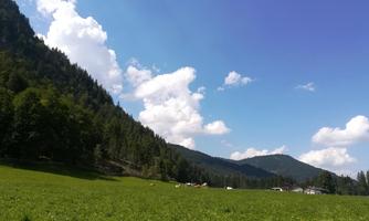 A green meadow surrounded by gentle hills and forested mountains. The sky is blue with some white clouds.