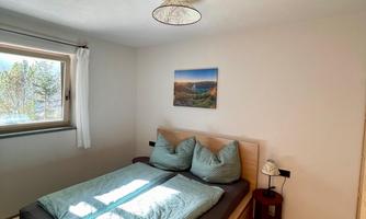 A cozy bedroom with a double bed and two pillows. A window offers a view of nature and a picture hangs on the wall.