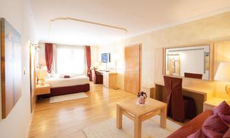 A spacious hotel room with a double bed, a desk, and comfortable seating. Large windows provide plenty of daylight and an inviting atmosphere.