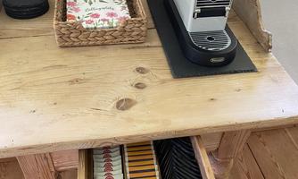 A wooden table with a modern coffee machine and a small basket with napkins. Drawer with coffee capsules and cookies underneath.