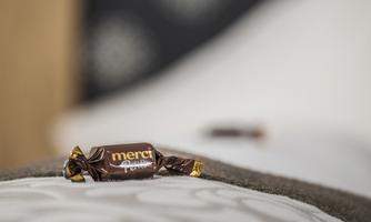 A single piece of chocolate from the brand "Merci" lies on a bedspread. The background is blurred and conveys a cozy atmosphere.