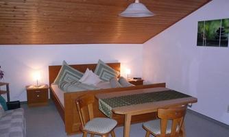 A cozy bedroom with a double bed and a seating area. The room has a wooden ceiling and is brightly lit.