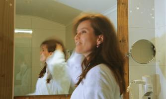 A woman in a bathrobe stands in front of a mirror. She smiles and has her hair slightly tousled.