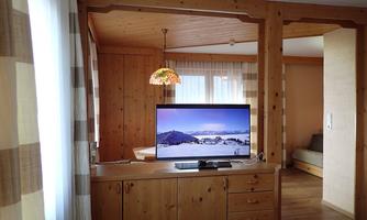 A cozy living space with wooden furniture and a large television. In the background, windows with curtains and a sofa are visible.