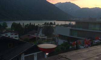 A cocktail glass stands on a wooden table overlooking a lake with mountains in the background. The sunset lights up the sky with soft colors.